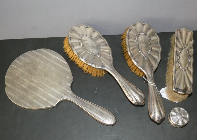 A set of vintage grooming tools including a handheld mirror, two hairbrushes, a clothes brush, and a small round container, all with ornate silver handles.